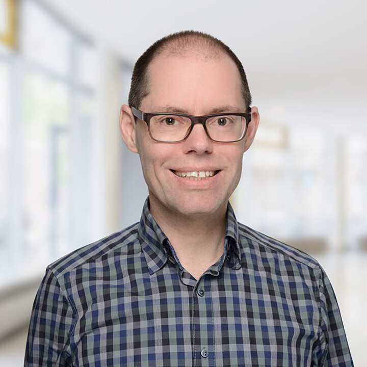 Portrait von Martin Peitz. Er trägt ein karriertes dunkelblau-schwarzes Hemd und lächelt.