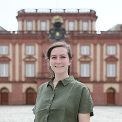 Tamara Schmitt hat braune Haare und trägt eine olivgrüne Bluse. Sie steht vor dem Schloss.