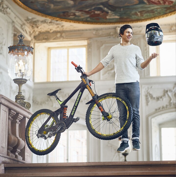 Sportlich aussehender Student steht schwebend über dem Boden in einem der Schlosssääle der Universität. In der rechten Hand hält er sein Mountainbike fest und über seiner linken Hand schwebt ein Motorradhelm.