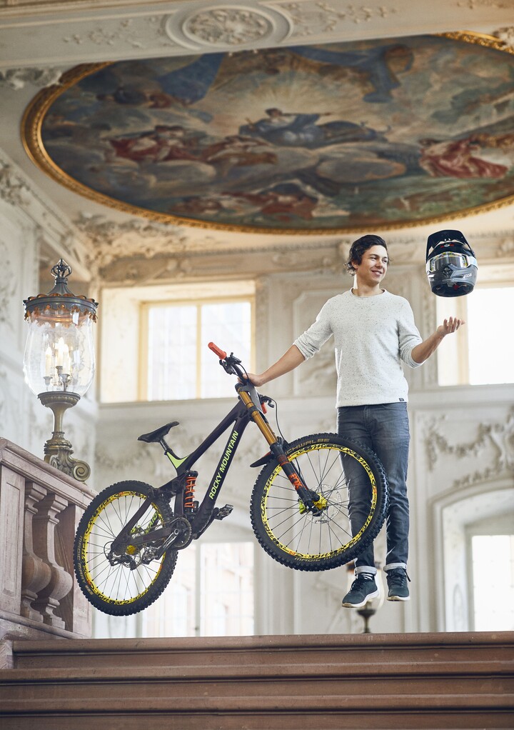 Sportlich aussehender Student steht schwebend über dem Boden in einem der Schlosssääle der Universität. In der rechten Hand hält er sein Mountainbike fest und über seiner linken Hand schwebt ein Motorradhelm.