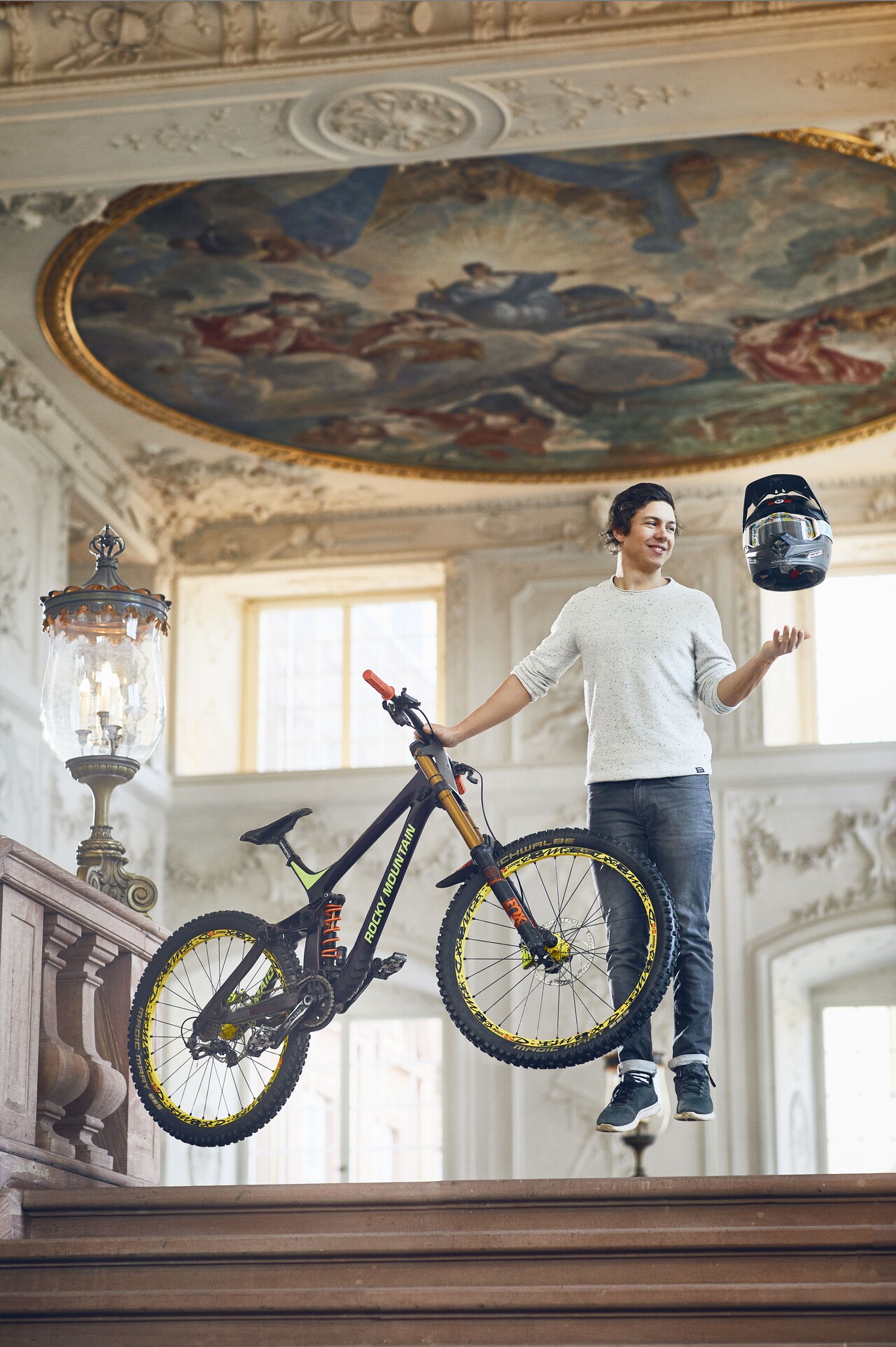 Sportlich aussehender Student steht schwebend über dem Boden in einem der Schlosssääle der Universität. In der rechten Hand hält er sein Mountainbike fest und über seiner linken Hand schwebt ein Motorradhelm.