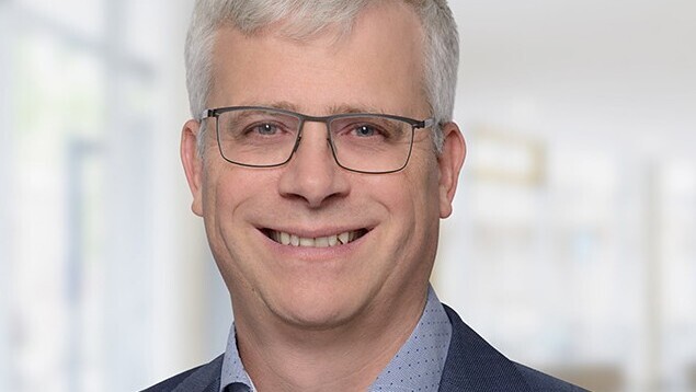 Portrait von Eckhard Janeba. Er trägt ein blaues Hemd sowie einen dunkelblauen Blazer und steht lächelönd vor einer Fensterfront.