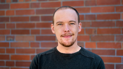 Jan Menzner. He is wearing a black sweater and is standing in front a brickwall.