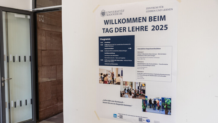 Eingang zu einem Veranstaltungsraum mit geöffneter Tür. An der Wand neben dem Eingang ist ein großes Poster mit dem Programm der Veranstaltung angebracht.