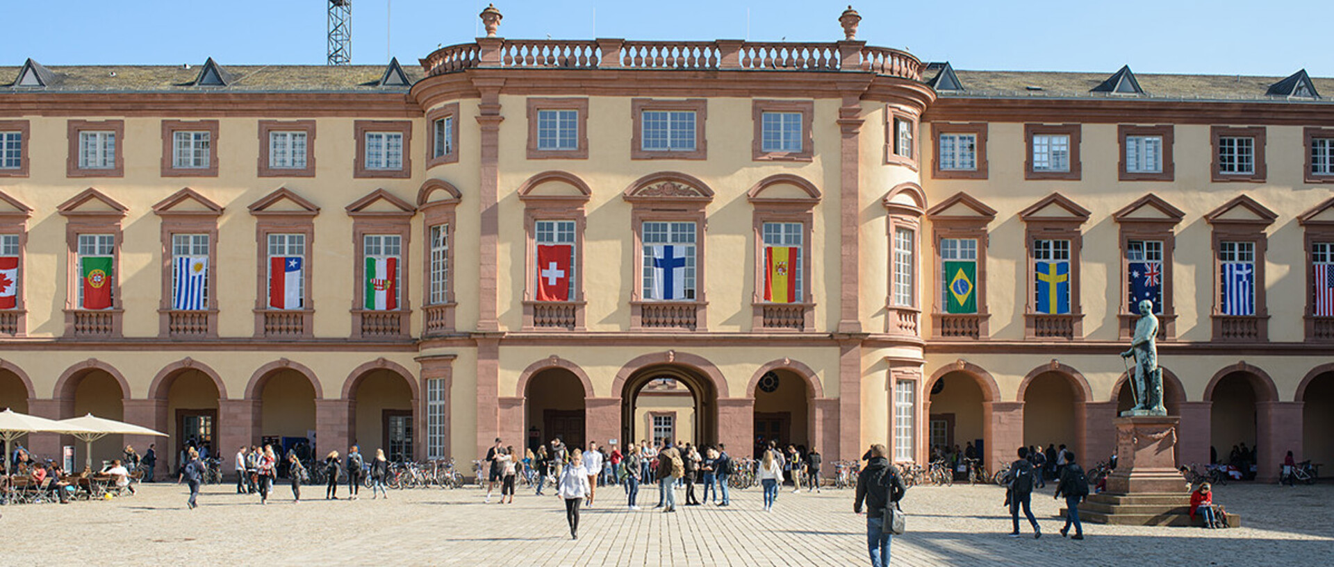 Die Fassade des Osflügel des Schlosses ist mit Länderflaggen dekoriert. Im Vordergrund überqueren Menschen den Ehrenhof.