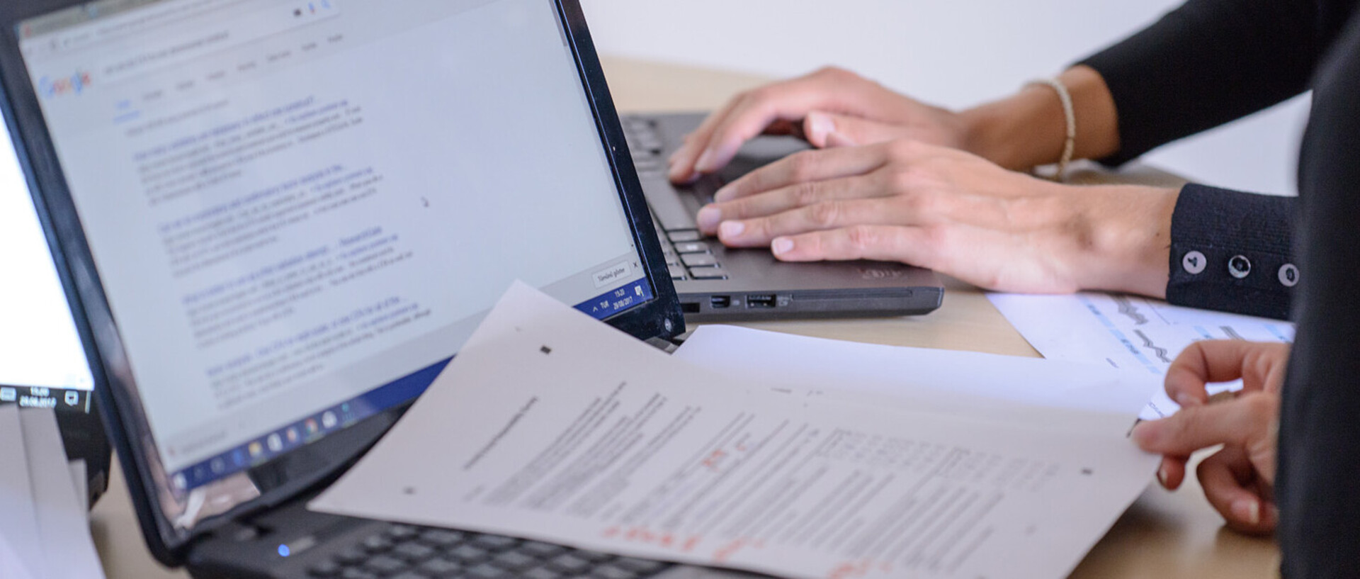 Zwei geöffnete Laptops auf einem Schreibtisch, Personen arbeiten mit Dokumenten.