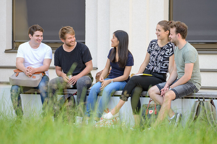Eine Gruppe von Studierenden sitzt in entspannter Atmosphäre draußen auf einer langen Bank