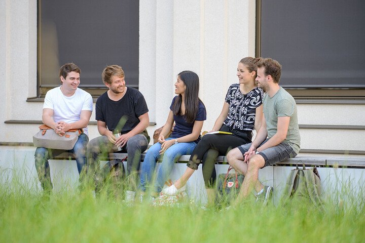 Eine Gruppe von Studierenden sitzt in entspannter Atmosphäre draußen auf einer langen Bank