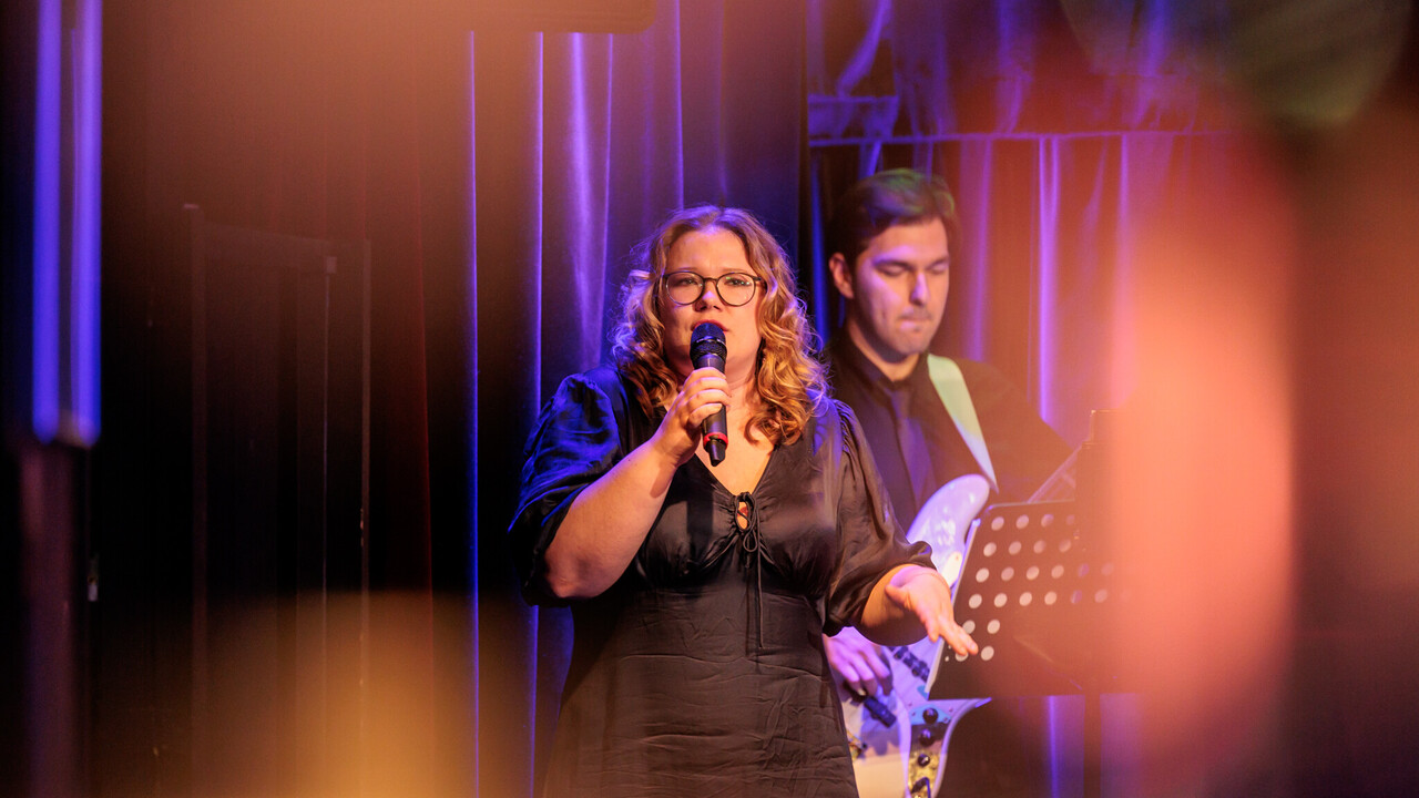 Eine Sängerin singt in ein Mikrofon. Im Hintergrund ist ein Gitarrist mit E-Gitarre zu sehen.