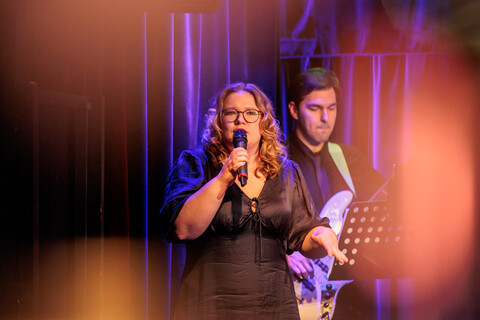 Eine Sängerin singt in ein Mikrofon. Im Hintergrund ist ein Gitarrist mit E-Gitarre zu sehen.