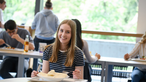 Eine Studentin sitzt mit Messer und Gabel in der Hand an einem Tisch in der Mensa und lächelt, auf ihrem Teller liegt ein Wrap.