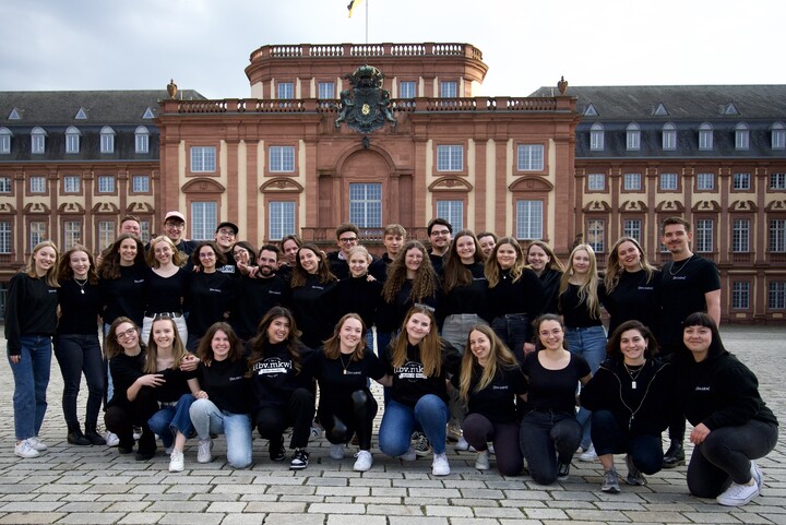 Die Fachschaft der Medien- und Kommunikationswissenschaften steht in schwarzer Fachschaftskleidung zusammen vor dem Schloss.