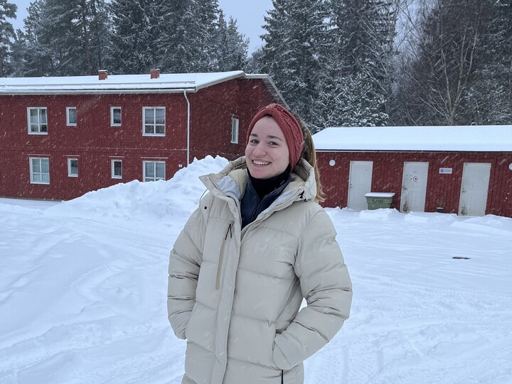 Lara trägt dcke Winterkleidung. Alles um sie ist eingeschneit, im Hintergrund sind rote Häuser und verschneite Tannen.