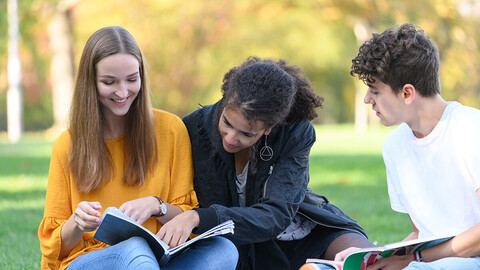 Drei Studierende sitzen mit ihren Büchern auf einer Wiese