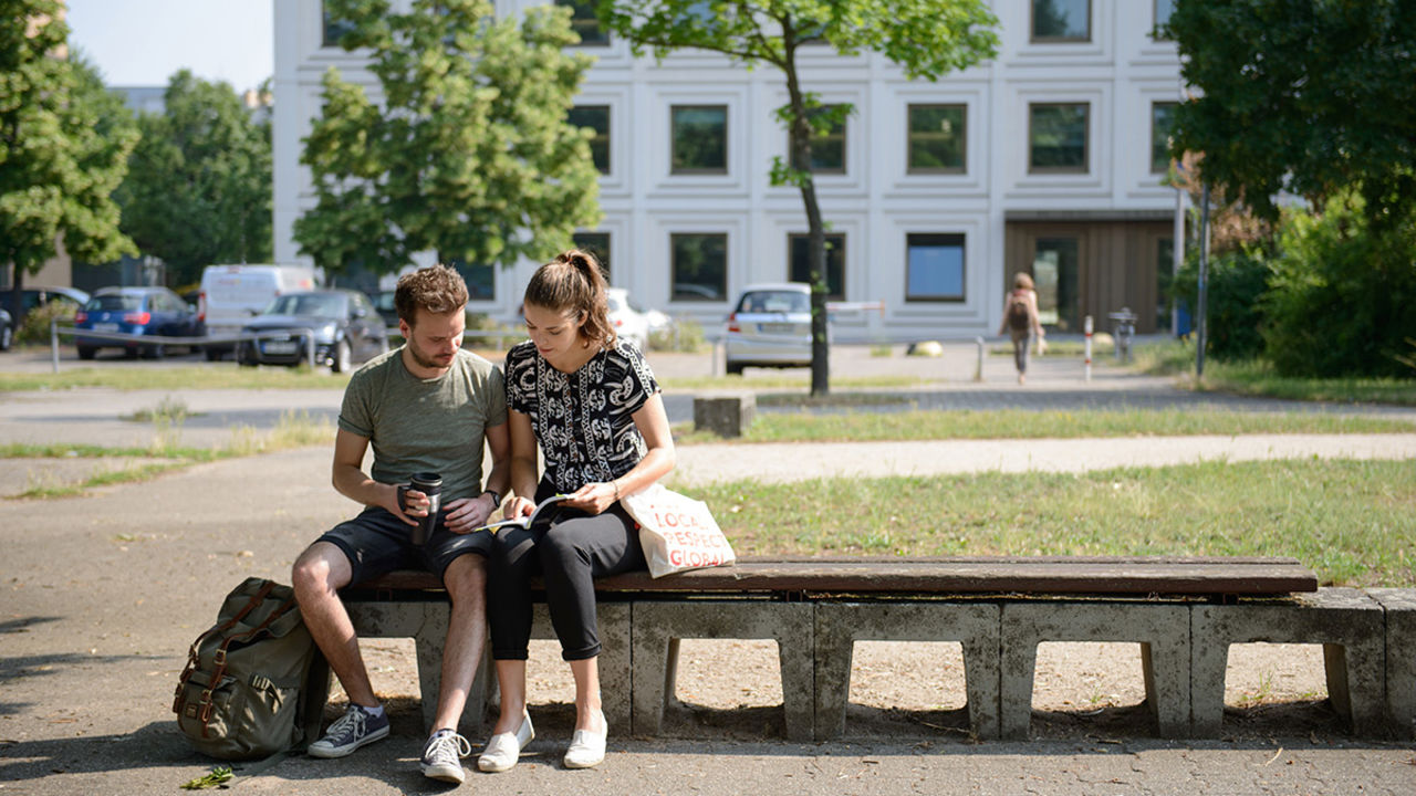 Eine Studentin und ein Student sitzen auf einer Bank vor dem Gebäude A6 der Universität Mannheim. Sie lesen gemeinsam in einem Buch.