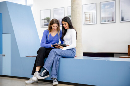 Zwei Stundentinnen sitzen vor einem Hörsaal und betrachten gemeinsam etwas auf einem Tablet.