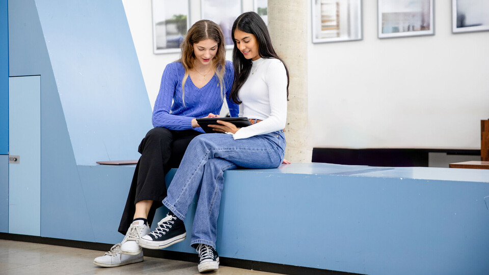 Zwei Stundentinnen sitzen vor einem Hörsaal und betrachten gemeinsam etwas auf einem Tablet.