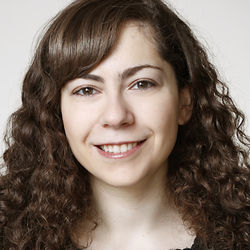 Anna Bruk hat lange braune Haare mit Locken. Sie trägt ein dunkles Oberteil und lächelt in die Kamera.