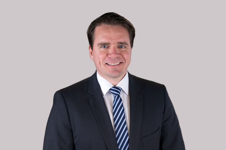 Prof. Höhle hat kurze dunkle Haare und trägt ein dunkles Jacket, ein weißes Hemd und eine gestreifte Krawatte.