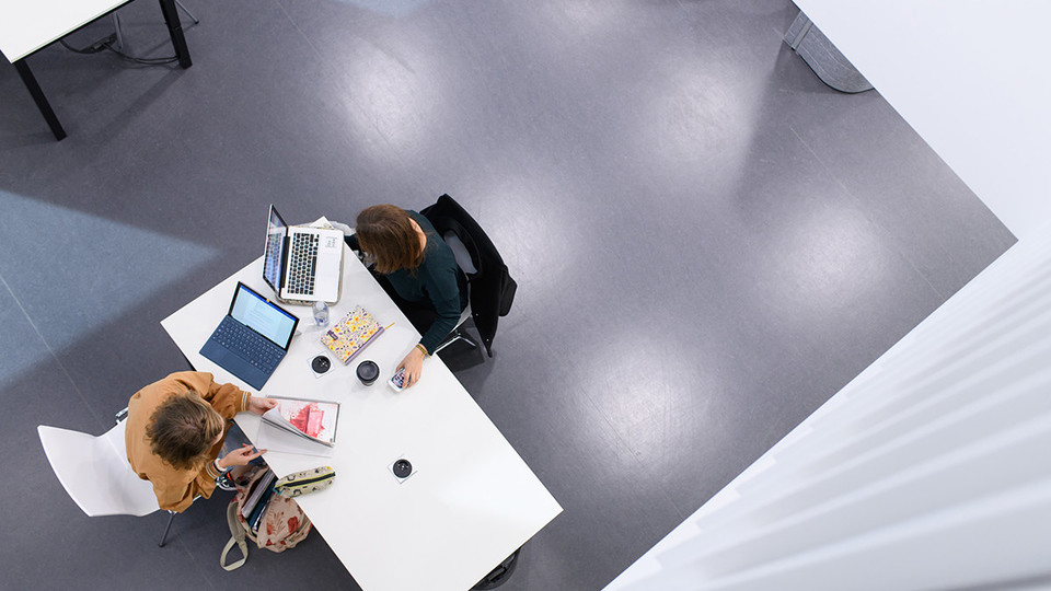 Vogelperspektive auf zwei Studentinnen, die mit ihren Laptops an einem weißen Tisch arbeiten.