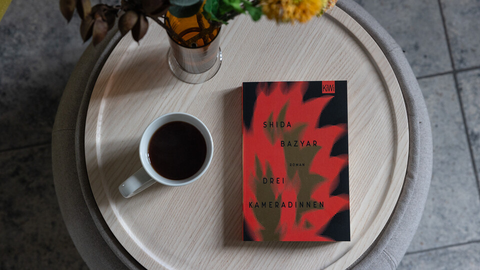 Das Buch "Drei Kameradinnen" liegt auf einem kleinen Tisch. Daneben ist eine Kaffeetasse und eine Blumenvase zu sehen.