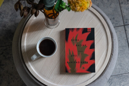 Das Buch "Drei Kameradinnen" liegt auf einem kleinen Tisch. Daneben ist eine Kaffeetasse und eine Blumenvase zu sehen.