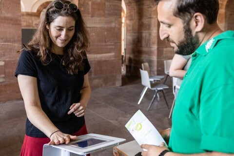 Gutgelaunte Frau macht Experiment und legt Hand auf einen digitalen Bildschirm. Neben ihr Mann mit AOK T-Shirt, welcher ihr dabei zuschaut.
