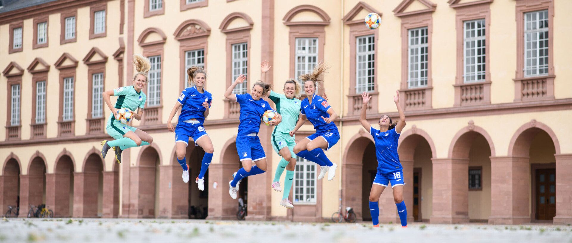 Spitzensport Fußball Sechs Fußballerinnen springen, in blauen Trikots gekleidet, vor dem Mannheimer Schloss in die Luft.