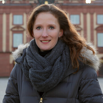 Eine Person trägt einen schwarzen Pollover, eine grau Jacke mit Fell sowie einen grauen Schal und steht auf dem Ehrenhof. Die Person heißt Karin Hoisl.