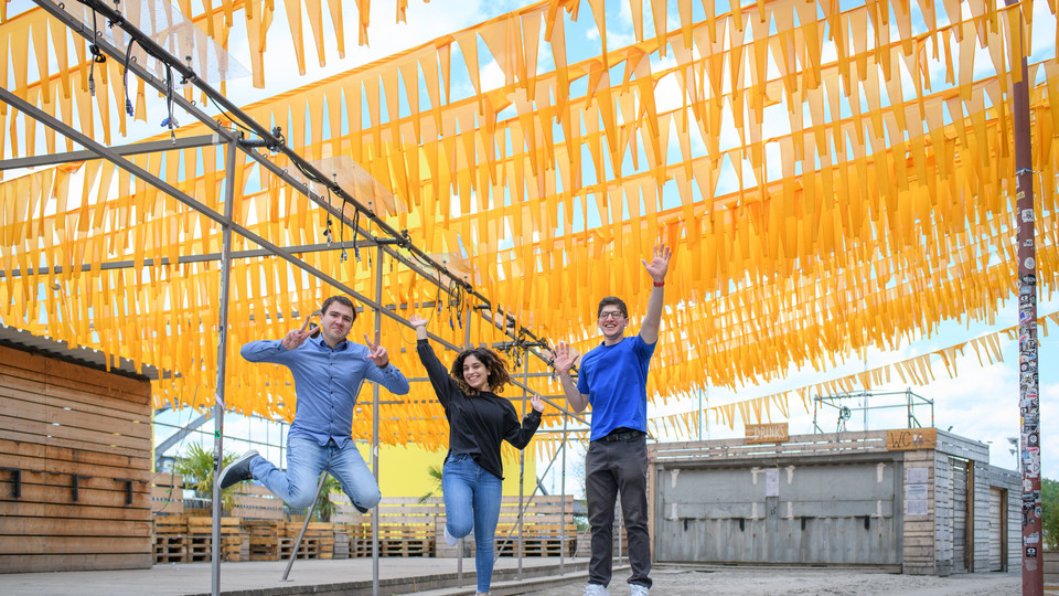 Drei Studierende springen jubelnd in die Luft. Kulisse ist das Mannheimer Hafengelände. Über ihnen sind gelbe Wimpel gespannt.
