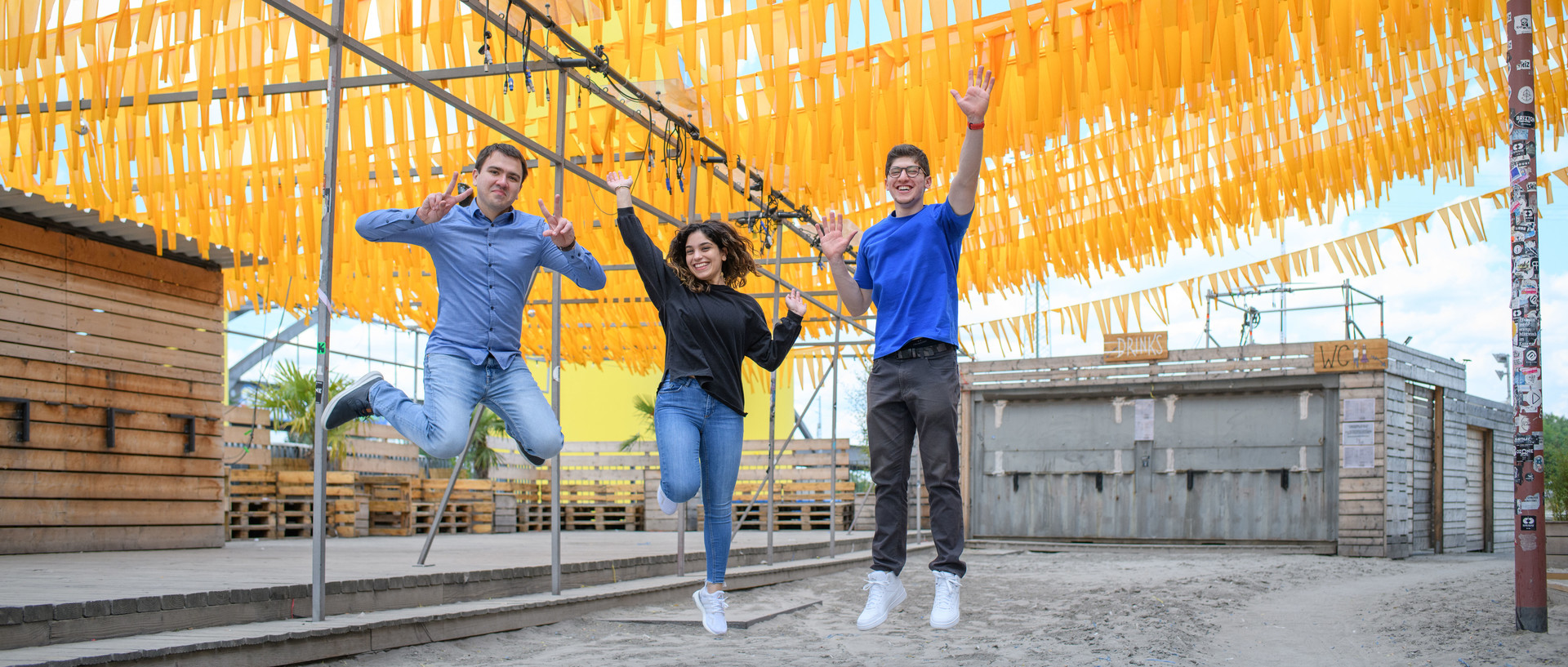 Drei Studierende springen jubelnd in die Luft. Über ihnen sind gelbe Wimpel. Link: Pressemeldung StudyCheck Award 2023