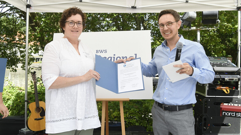 Zwei Menschen stehen im Freien unter einem Pavillon und halten beide eine Urkunde. Sie schauen in die Kamera.