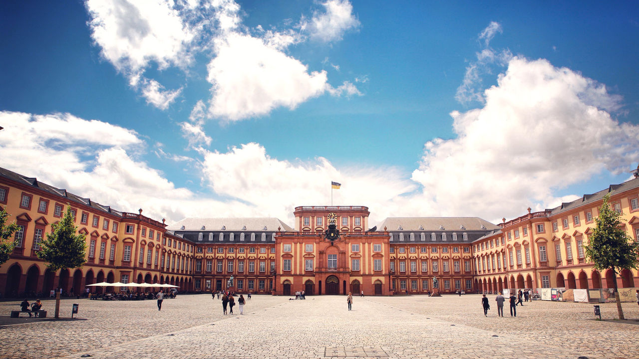 Das Barock-Schloss und der Ehrenhof der Universität Mannheim unter strahlend blauem Himmel. Das Schloss ist von unzähligen Fenstern, rotem Sandstein und einer gelben Fassade geprägt.