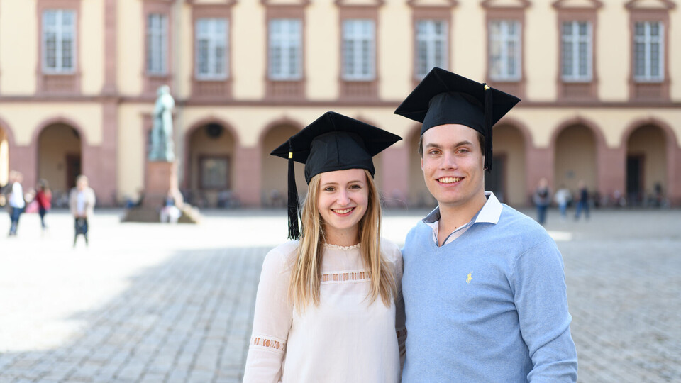Eine Studienabsolventin und ein Studienabsolvent stehen auf dem Ehrenhof Zwei Studierende mit Hüten vor dem Schloss
