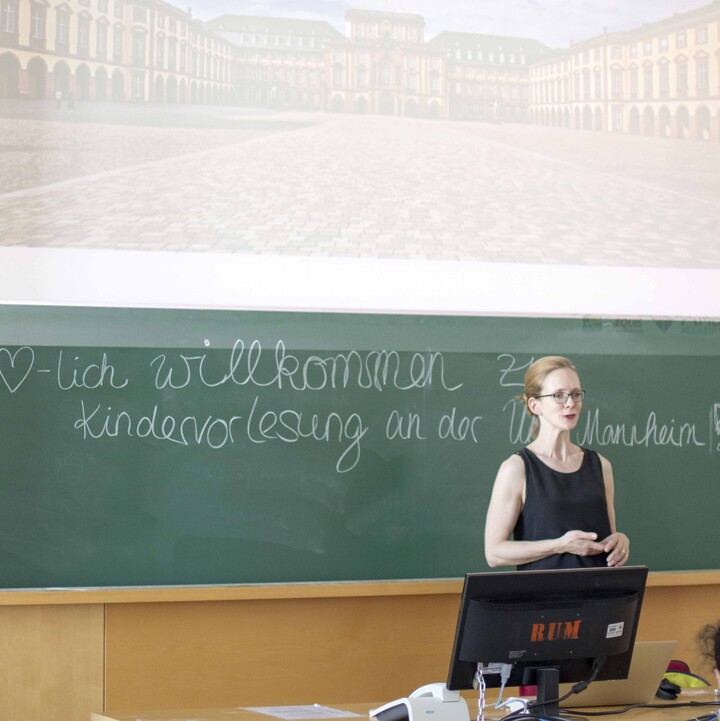 Eine Dozentin steht vor einer Tafel mit der Aufschrift "Herzlich Willkommen zur Kindervorlesung an der Uni Mannheim". Darüber wird eine Folie mit der Aufschrift "Was ist ein Unternehmen?" gezeigt. 