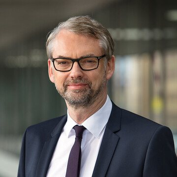 Micheal Strugl hat kurze graue Haare. Er trägt eine dunkle Brille, ein weißes Hemd, eine braune Krawatte und einen dunklen Anzug darüber