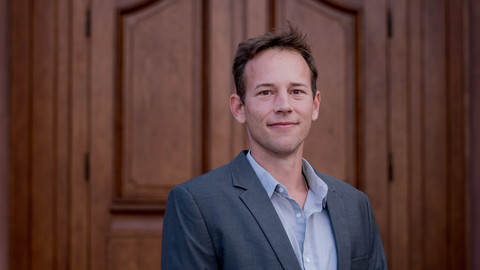 Samuel Wissel. He is wearing a grey suit and is standing in front of a brown door.