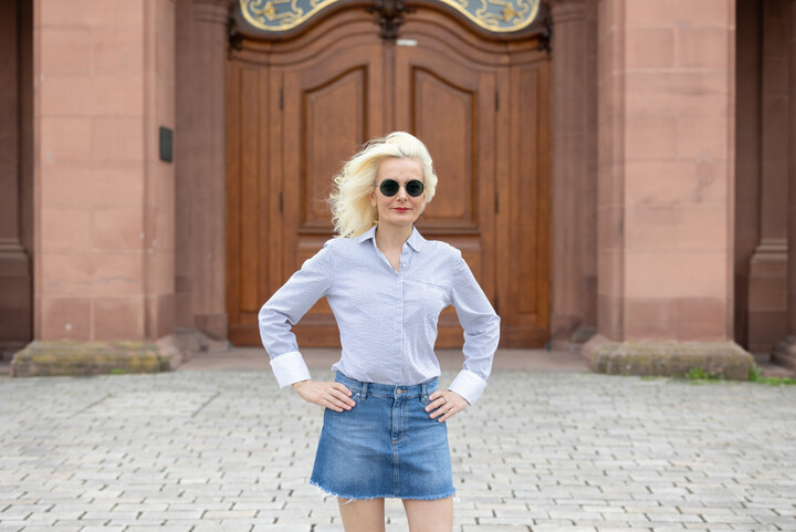 Tülay Yavuz from Türkiye stands in the Ehrenhof with her arms on her hips. She wears dark sunglasses and her dyed blond hair flutters in the wind. She looks very confident, wearing red lipstick, a denim skirt and a light blue blouse. Link: MyUniMA story August 2023