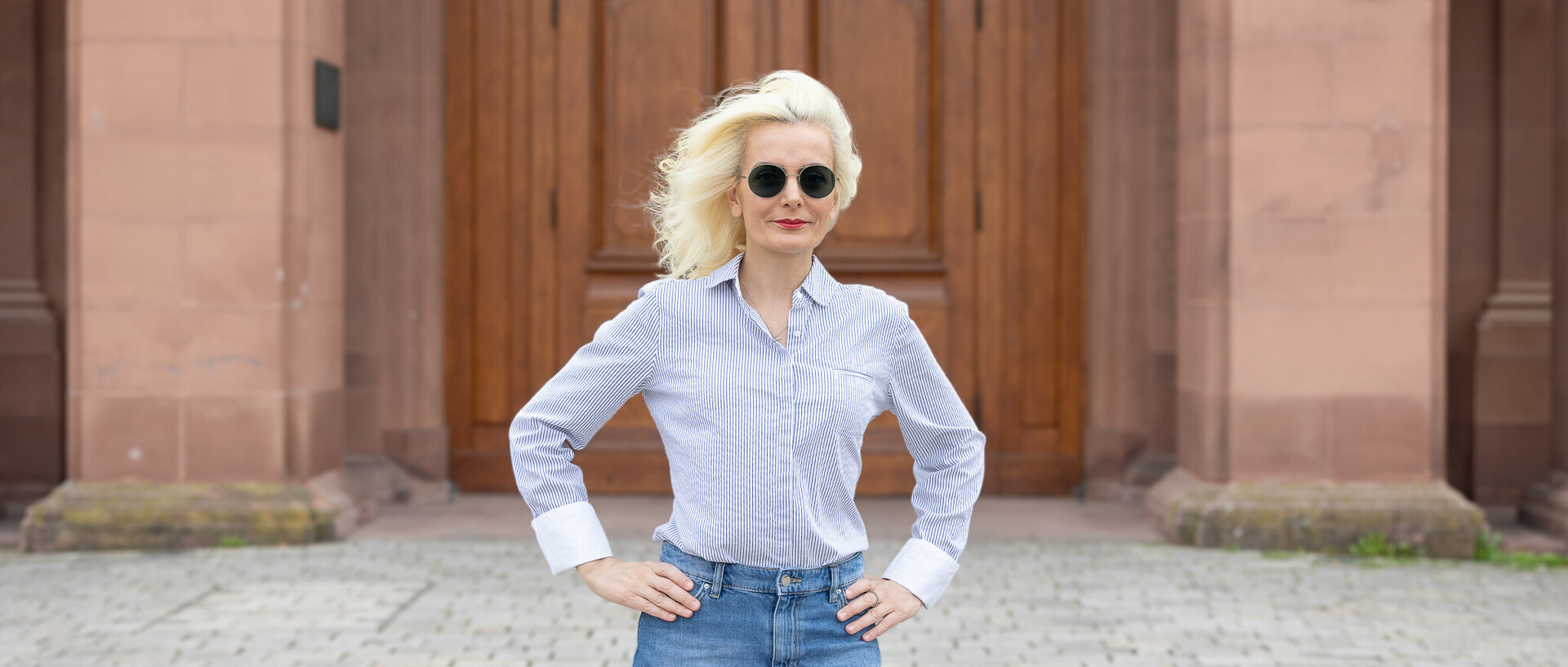 Eine Person trägt ein weiß-hellblau gestreiftes Hemd sowie einen Jeansrock und steht vor einer braunen Schlosstüre. Die Person heißt Tülay Yavuz.