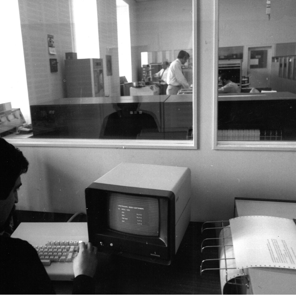 Auf einem alten Schwarz-Weiß-Foto sehen wir zwei Männer vor sehr alter Computer Hardware. Sie befinden sich jeweils in einem Büro, die durch eine Glasscheibe getrennt sind.