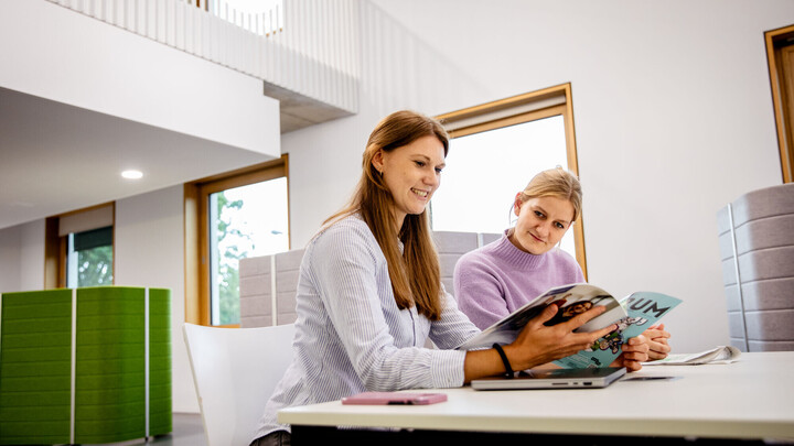 Zwei weiblich gelesene Studierende sitzen nebeneinander an einem Tisch in der Bibliothek und lesen lächelnd in einem Universitätsmagazin.