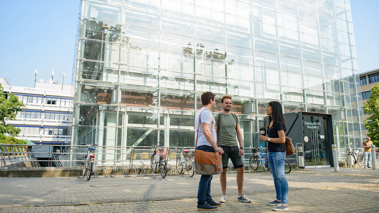 Eine Studentin und zwei Studenten unterhalten sich vor dem gläsernen Gebäude A5 der Universität Mannheim. Sie tragen T-Shirts und Taschen.