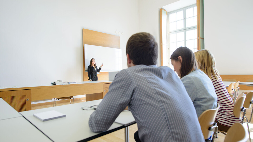 Drei Studierende sitzen in einem Seminarraum und blicken zu einer Dozentin, die vor ihnen an der Tafel steht. 