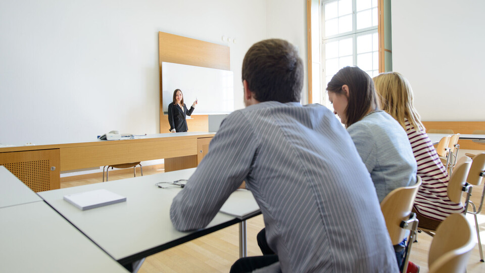 Drei Studierende sitzen in einem Seminarraum und blicken zu einer Dozentin, die vor ihnen an der Tafel steht.
