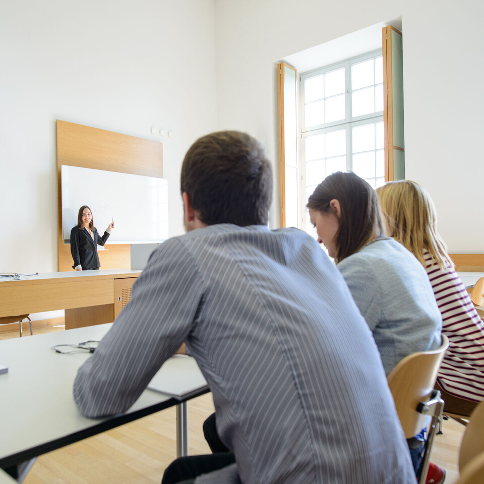 Drei Studierende sitzen in einem Seminarraum und blicken zu einer Dozentin, die vor ihnen an der Tafel steht. 