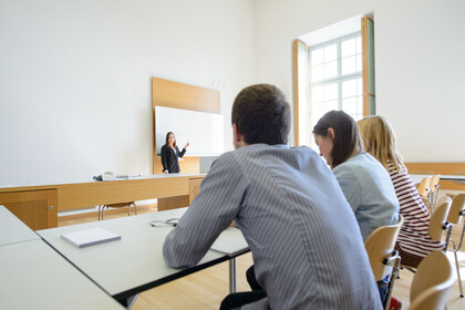 Drei Studierende sitzen in einem Seminarraum und blicken zu einer Dozentin, die vor ihnen an der Tafel steht. 