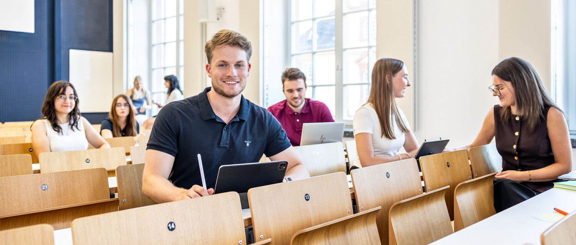 Junger Mann mit blauem T-Shirt und blondem Haar sitzt in einem Hörsaal und schaut in die Kamera.