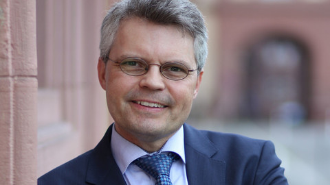 Eine lächelnde Person trägt ein flieder farbenes Hemd, eine gemusterte Krawatte sowie einen dunkelblauen Blazern und steht angelehnt an eine Wand. Die Person heißt Ralf Müller-Terpitz.