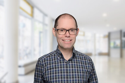 Portrait von Martin Peitz- Er trägt ein schwarz-blau karriertes Hemd und steht lächelnd vor einer Fensterfront.