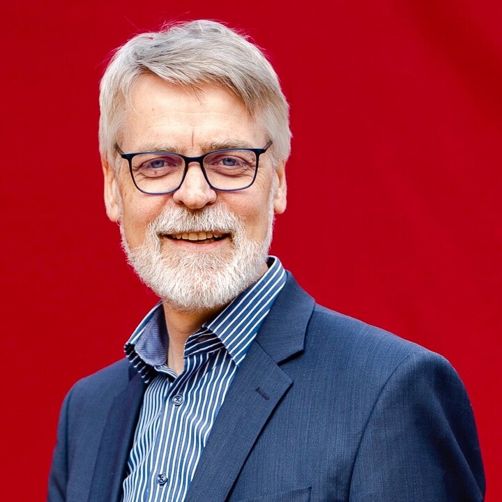 Hartmut Wessler vor einem roten Hintergrund. Er trägt eine Brille und einen dunkelblauen Anzug.